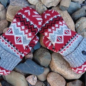May include: A pair of knit mittens featuring a red, white, and gray geometric fair isle pattern. The mittens have a gray ribbed cuff with a single black button detail on the front of each cuff, finished with a red and white zigzag border.
