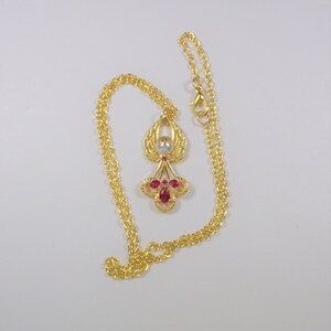 May include: A gold chain necklace with a pendant featuring a stylized angel with red and white gemstones.
