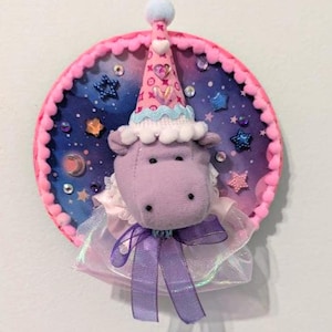 May include: A plush hippo toy with a party hat and bow, set against a circular backdrop. The backdrop has a starry night sky design with pink trim and decorative gems. The hippo is lavender with a white collar and a purple ribbon.