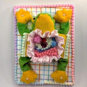 May include: A whimsical plush frog art piece. The frog is primarily green and yellow, with a pink ruffled chest and a multi-colored interior. The background is a checkered pattern with a pink pom-pom border. The frog's feet and hands have pearl accents.