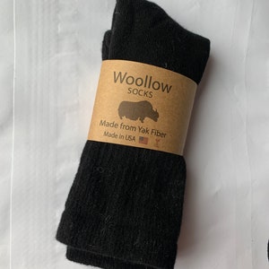 Pode incluir: Meias pretas de fibra de iaque, embrulhadas com uma etiqueta de papel castanho. A etiqueta diz "Woollow Socks" com uma ilustração de iaque e "Made in USA". As meias estão dobradas e parecem macias.