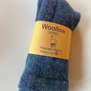 May include: Blue wool socks with a tan paper band. The band reads "Woollow SOCKS" and features an alpaca illustration. The socks are made from Huacaya Alpaca wool. The label also states "Made in USA".