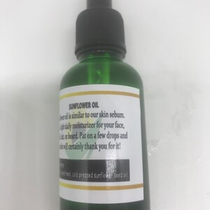 May include: A green glass bottle with a black dropper top. The label reads "SUNFLOWER OIL" and describes it as a daily moisturizer. The label also lists the ingredients as refined, cold pressed sunflower seed oil.
