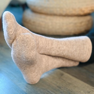 Könnte beinhalten: Nahaufnahme von zwei hellbraunen Socken, ein Fuß ruht auf dem anderen. Die Socken sind aus einem weichen, strukturierten Material gefertigt. Der Hintergrund umfasst einen Holzboden und eine verschwommene Ansicht eines geflochtenen Korbs und eines Stuhls.