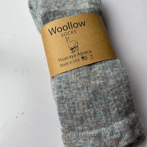 May include: A pair of gray and blue speckled socks, wrapped with a brown paper band. The band features the text "Woollow SOCKS", a drawing of an alpaca, and the words "Huacaya Alpaca" and "Made in USA".
