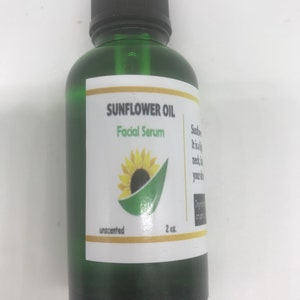 May include: A green glass bottle of Sunflower Oil Facial Serum. The label features a sunflower illustration and the text "unscented" and "2 oz.". The bottle has a black dropper top.
