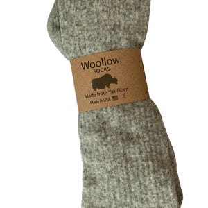 May include: A pair of light gray Woollow socks made from yak fiber. The socks are folded and held together with a brown paper band that reads "Woollow Socks" with a yak silhouette and "Made from Yak Fiber Made in USA".