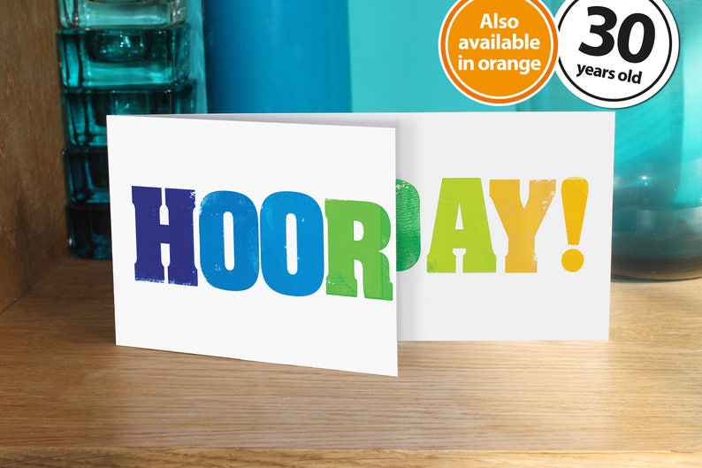 May include: Two white greeting cards with the word "HOORAY!" printed in rainbow colours. The cards are on a wooden surface.