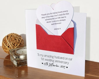 May include: A white card with a red envelope attached to it. The card has a message that says "To my amazing husband on our 1st wedding anniversary 17th September 2019". The envelope has a heart-shaped note that says "Thank you for being there every step of the way. Can't wait for the adventures our life has in store! Love you always and forever! Jenny x"