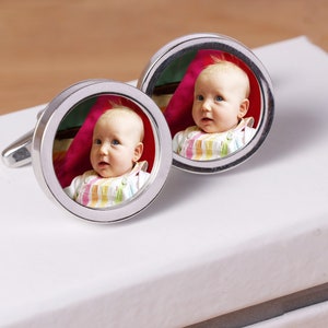 May include: A pair of silver cufflinks with a round photo insert featuring a baby in a striped outfit. The baby is looking at the camera and smiling.