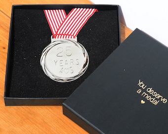 May include: A silver medal with the inscription "25 YEARS 406" and a red and white striped ribbon, presented in a black box. The box is open, revealing the medal and the text "You deserve a medal".