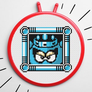 Może przedstawiać: Pikselowana postać w kolorze niebieskim i białym z koroną i złymi oczami jest wyszyta na białym płótnie w czerwonym obręczy do haftu.