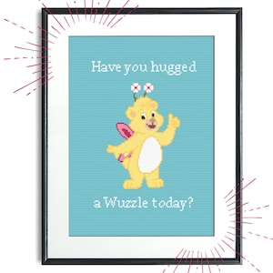 Könnte beinhalten: Ein gerahmtes Kreuzstich-Design mit einem gelben Bären mit rosa Flügeln und weißen Blumen auf dem Kopf. Der Bär lächelt und winkt. Der Text "Have you hugged a Wuzzle today?" steht unter dem Bären.
