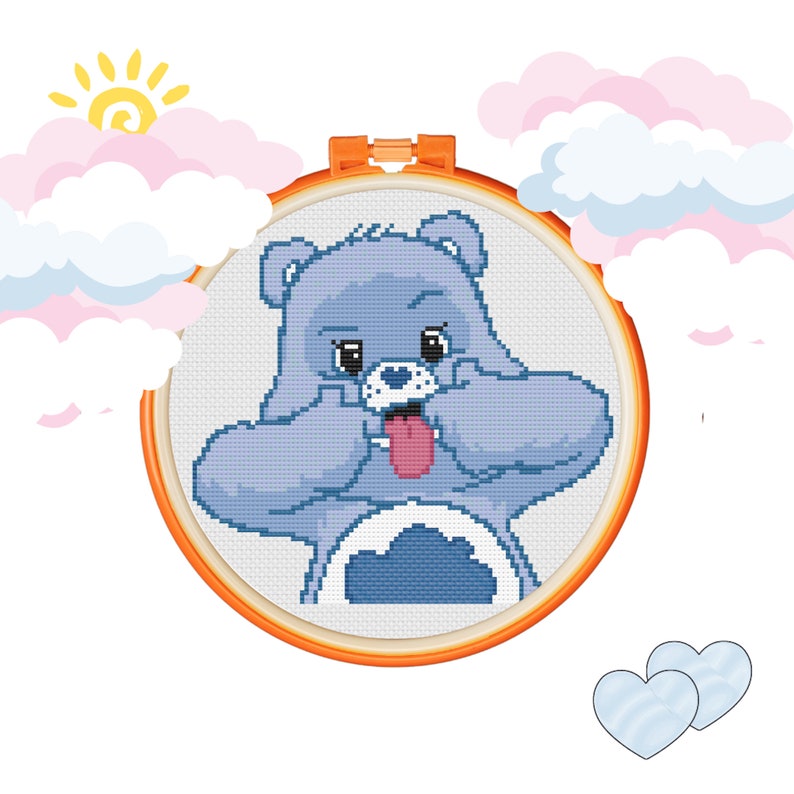 May include: A blue Care Bear with a tongue sticking out, holding its paws up to its face, in a circular embroidery hoop. The Care Bear is against a white background with pink and blue clouds.