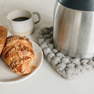 Peut inclure: Une tasse en céramique blanche remplie de café se trouve à côté de deux croissants aux amandes sur une assiette blanche mouchetée. Une carafe à café argentée repose sur un dessous de plat en tricot gris.