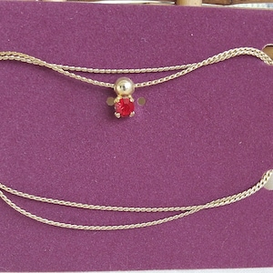 May include: A gold chain necklace with a red gemstone pendant. The necklace is displayed on a purple velvet background.