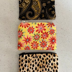May include: Three small zippered pouches with different patterns. The top pouch has a black and gold paisley pattern. The middle pouch has a yellow background with red and pink flowers. The bottom pouch has a brown and black leopard print.