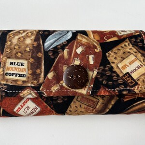 May include: A brown fabric zippered pouch with a coffee bean print. The pouch has a brown coconut button closure and features various coffee brands including "Blue Mountain Coffee" and "100% Arabica Mocha".