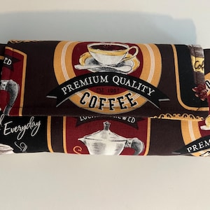 May include: A brown fabric clutch with a coffee theme. The clutch has a gold and red design with the words "Premium Quality Coffee" and "Served Everyday".
