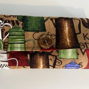 May include: A brown fabric wallet with a coffee theme. The wallet has a button closure and features a repeating pattern of coffee pots, coffee beans, and the words "Cafe" and "Kaffee Spritz".