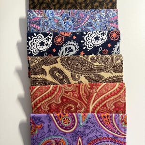 May include: A stack of six fabric wallets with paisley patterns. The wallets display a variety of colors, including brown, blue, navy, tan, red, and purple. Each wallet has a unique paisley design.