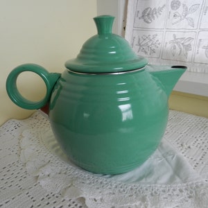 May include: A green enamel teapot with a silver rim and a lid. The teapot is sitting on a white crocheted doily.