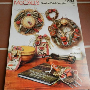 May include: A McCall's Crafts pattern for making a variety of garden-themed crafts, including wreaths, a bowl, and a book. The pattern is titled "Garden Patch Veggies" and has the number 15084. The image shows a variety of finished crafts, including a straw hat wreath, a wreath with potted plants, a bowl with a red ribbon, and a book titled "The Impressionist Garden".