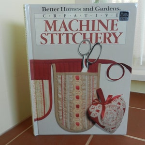 May include: A book titled "Better Homes and Gardens Creative Machine Stitchery" with a red, white, and beige fabric pocket on the cover. The pocket contains a pair of scissors and a heart-shaped pincushion.