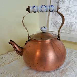 May include: A copper kettle with a blue and white porcelain handle and lid. The kettle is sitting on a white crocheted tablecloth.