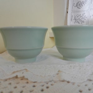 May include: Two light green ceramic bowls with a slightly rounded shape. The bowls are sitting on a white lace tablecloth.