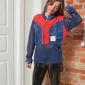 May include: A woman wearing a blue and red jacket with a zipper and a mesh panel. She is also wearing black leggings and red ankle boots.
