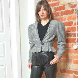 May include: A woman wearing a black and white striped blazer with puffed sleeves and black leather pants. She is standing in front of a brick wall and wearing black heeled boots.