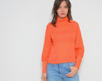 May include: A woman wearing a bright orange turtleneck sweater with long sleeves and blue jeans with a patterned stripe down the side. She is wearing purple ankle boots.