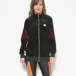 May include: A black velvet zip-up jacket with maroon accents on the sleeves. The jacket has a gold zipper and a small circular logo on the chest. The model is wearing black leggings with colorful stripes and orange heels.