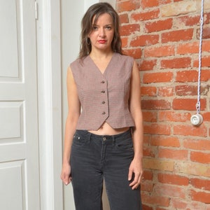 May include: A woman wearing a brown and white plaid vest with a black button closure. She is also wearing black jeans and black shoes.