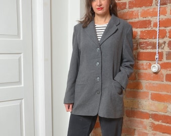May include: A woman wearing a gray blazer over a striped shirt and black pants. She is standing in front of a brick wall.