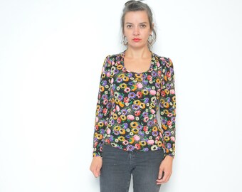 May include: A woman wearing a black long-sleeved blouse with a colorful floral print and dark gray jeans with slits at the bottom. She is wearing nude heels.