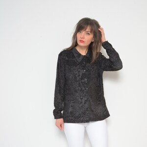 May include: A black long-sleeved shirt with a floral pattern and a collared neckline. The shirt is button-up and has a relaxed fit. The model is wearing white pants and black ankle boots.