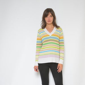 Puede incluir: Una mujer con un suéter de cuello en V a rayas de colores y pantalones negros. El suéter presenta rayas horizontales en tonos de amarillo, verde, azul y rosa. Los pantalones son negros y tienen una abertura en el tobillo.