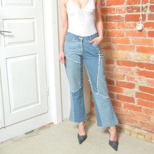 Könnte beinhalten: Eine Frau trägt einen weißen Spitzenbody und eine blaue Jeans mit Patchwork-Details. Sie steht vor einer Backsteinwand und einer weißen Tür.