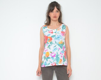 May include: A white sleeveless top with a floral print featuring pink, orange, and blue flowers. The top has a button closure at the neckline. The model is wearing brown pants and black heeled sandals.