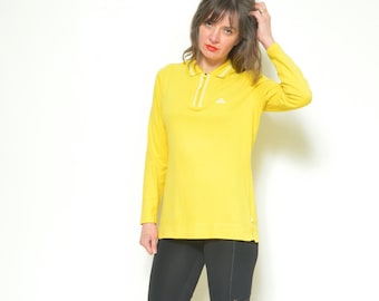 May include: A woman wearing a yellow long-sleeved collared shirt with a white zip and a small logo on the chest. She is wearing black leggings and yellow knee-high boots.
