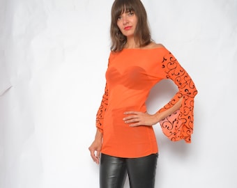 May include: A woman wearing a bright orange top with black floral detailing on the sleeves and black leather pants. She is wearing blue denim heeled mules.