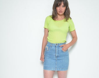 Puede incluir: Una mujer con una camiseta verde lima de manga corta y una minifalda vaquera azul claro. También lleva tacones altos naranjas. El conjunto se presenta sobre un fondo blanco.