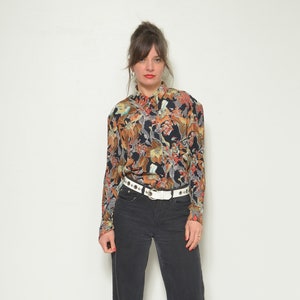 Può includere: Una donna indossa una camicia a maniche lunghe con stampa floreale nera e marrone con una cintura bianca con borchie e jeans neri a gamba larga.