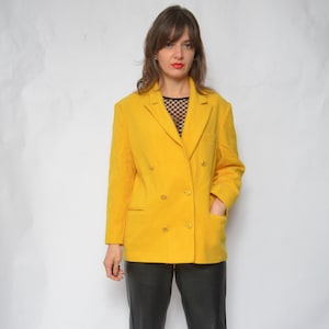 Peut inclure: Un blazer jaune vif à double boutonnage avec des boutons dorés. Le blazer est porté sur un haut en maille noire et un pantalon en cuir noir.