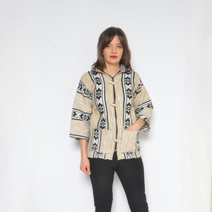 Peut inclure: Veste beige à motifs géométriques noirs et blancs, trois fermetures à bascule en bois et deux poches avant. La veste a des manches trois-quarts et est portée avec un pantalon noir et des talons bleus.