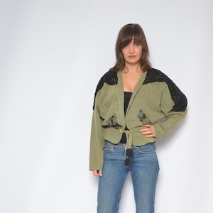 May include: A woman wearing a green and black jacket with a tie belt, blue jeans, and brown woven heeled mules.