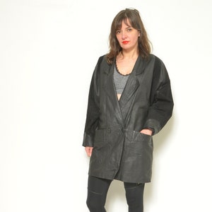 May include: A black leather jacket with a long, double-breasted design. The jacket has a collar and two pockets. The model is wearing black leggings and black boots.
