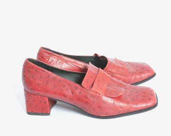 Könnte beinhalten: Ein Paar rote Loafer mit Absatz und Straußenmuster. Die Loafer haben eine quadratische Spitze und ein kleines Quasten-Detail.